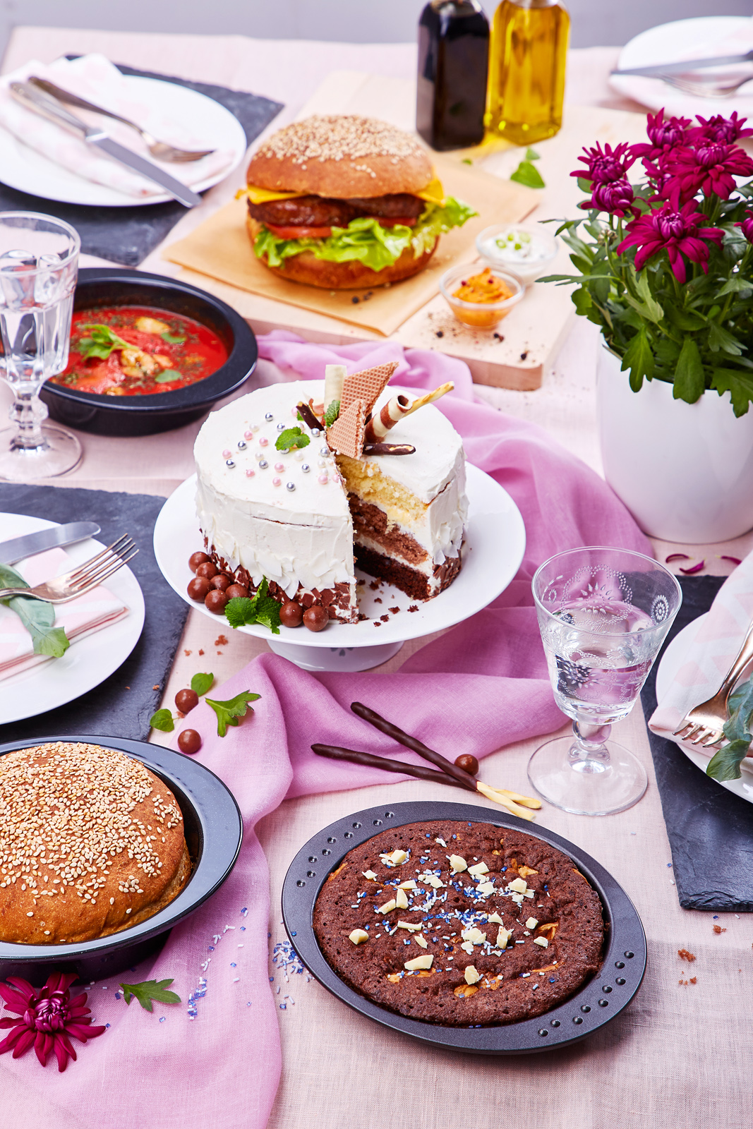 Servierformen mit Burger, Kuchen und weiteren Gerichten in einem farbig inszenierten Food-Arrangement