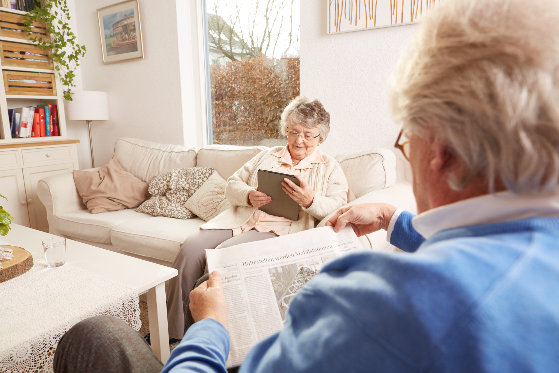 Seniorin nutzt ein Tablet im Wohnzimmer, während ein Senior Zeitung liest