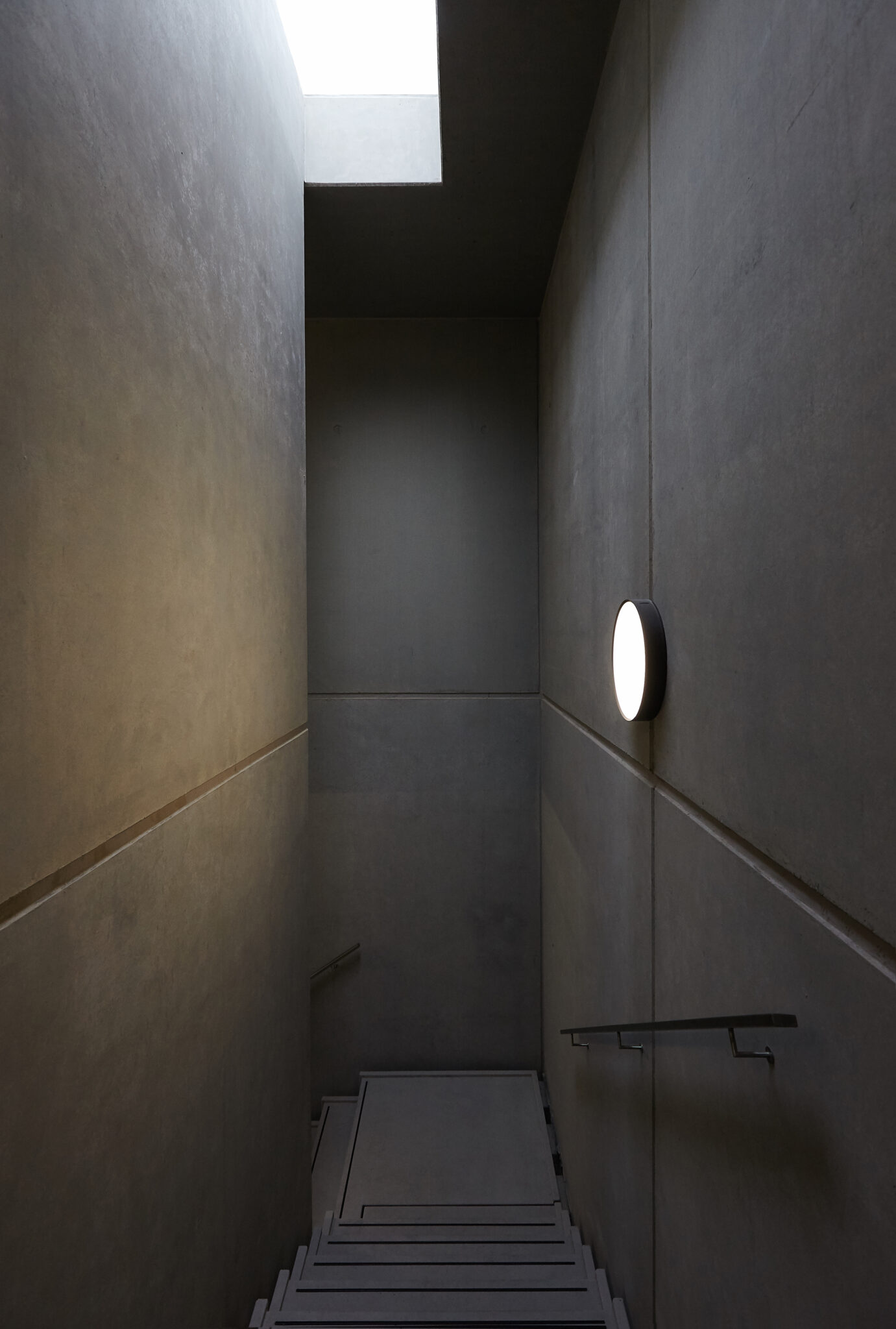 Schmales Treppenhaus aus Sichtbeton mit Oberlicht und runder Wandleuchte