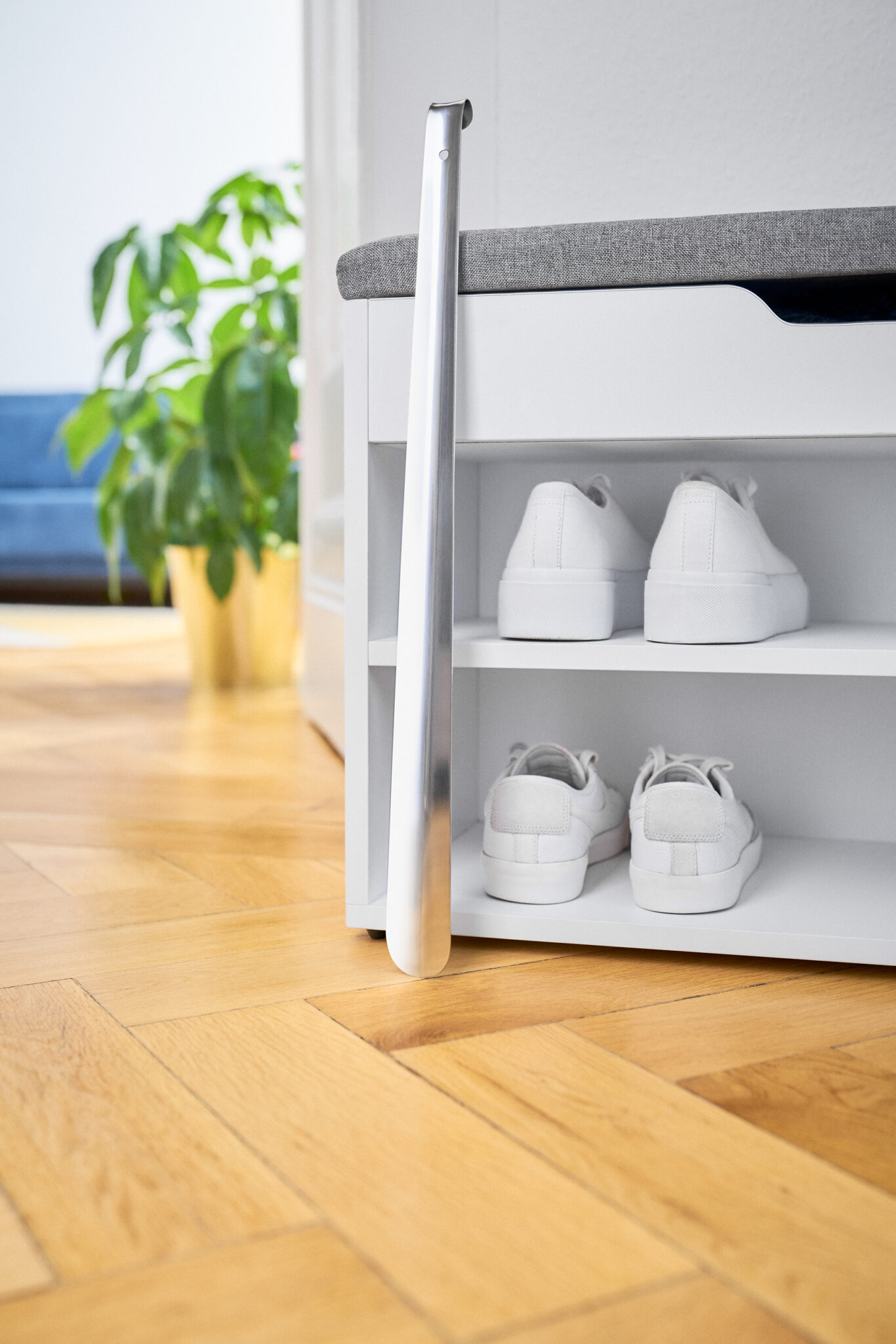 Langer Edelstahl-Schuhlöffel lehnt an einer weißen Schuhbank mit weißen Sneakern